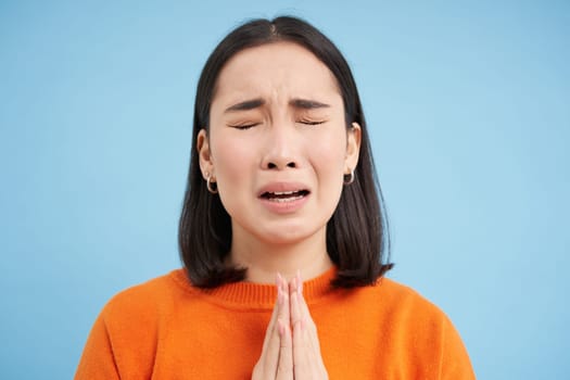 Close up of sad young asian woman, holds hands in pray, begging, pleading for help, stands over blue background