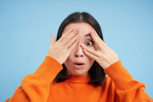 Close up portrait of korean girl waits for surprise, shuts her eyes and peeks through fingers with excited smile, stands over blue background