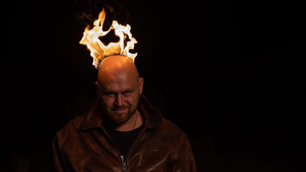 A bald man in a leather jacket with a burning head grins.
