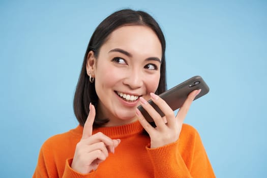 Portrait of korean woman records voice message on smartphone app, talks on speaker, translates her speech on mobile app, blue background