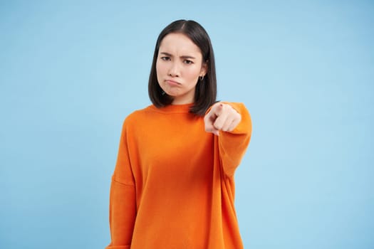 Angry frowning asian woman pointing finger, accusing, blaming someone, being disappointed, standing over blue background