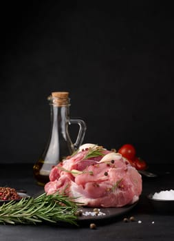 A piece of pork ham on a black board and spices olive oil, salt, rosemary branch and pepper