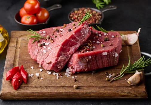 Raw piece of beef with spices pepper, rosemary sprig, salt and olive oil on a wooden board, black background