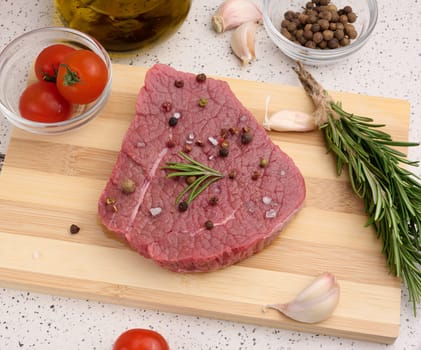 Raw piece of beef with spices pepper, rosemary sprig, salt and olive oil on a wooden board, top view