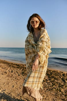 a woman wrapped in a plaid stands on the seashore in bright sunglasses