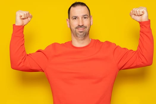 Bearded Hispanic man in his 40s wearing a red sweater showing off his pathetic biceps and pitiful shape while smiling proudly at the camera, isolated over yellow background.