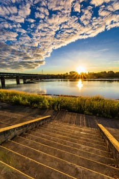 A beautiful sunrise over a wide river next to a long green bridge seen from the steps of the city promenade