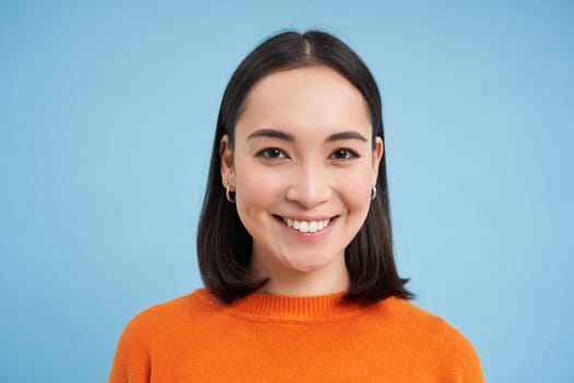 Close up portrait of asian girl with perfect healthy smile and natural beautiful face, looks happy at camera, stands over blue background