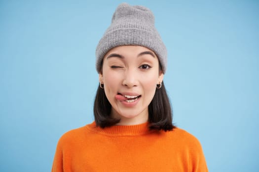 Stylish asian woman in beanie, shows tongue and winks at you, looks carefree, stands in orange sweatshirt over blue background