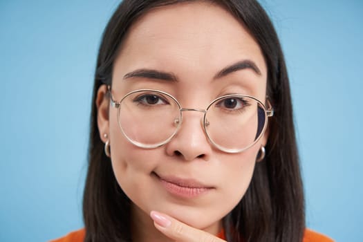 Close up portrait of asian woman looks intrigued, wears glasses, squints thoughtful, thinking, making assumption, standing over blue background