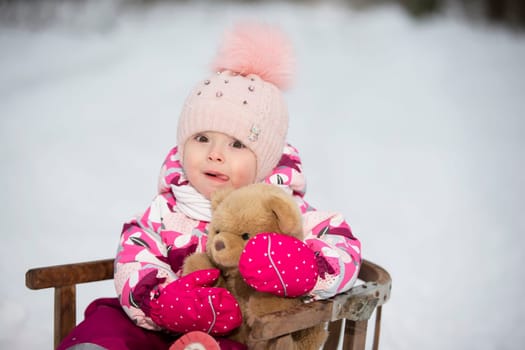 A little girl sits in a wooden sled with a teddy bear in winter.