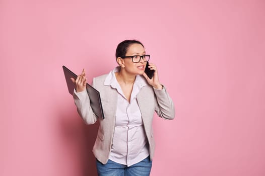 Multitasking overworked busy pregnant woman holding laptop and arguing while talking on mobile phone on pink backdrop