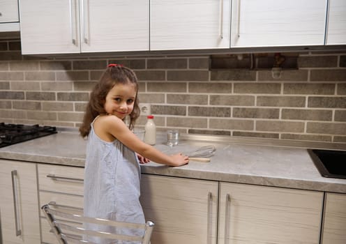 Adorable Caucasian baby girl standing on a chair by kitcheb countertop and smiles looking at camera through her shoulders