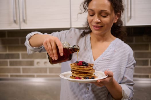 Soft focus on the hands of a beautiful female chef, charming pretty woman pouring maple syrup on a stack of homemade pancakes garnished with berries and cashew nuts. Shrove Tuesday concept, food art