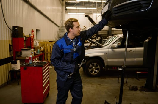 Handsome young mechanic in professional uniform, holding a wrench and working in auto service with lifted vehicle. Car repair and maintenance.