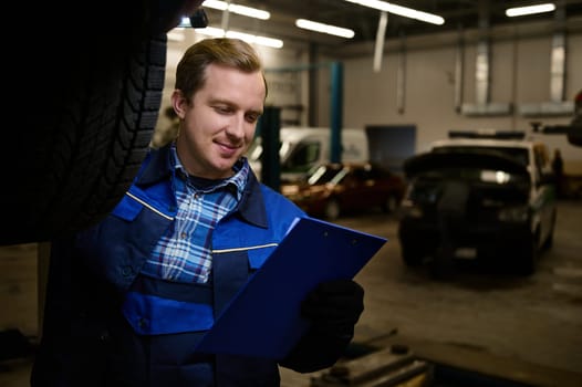 Professional portrait of a handsome Caucasian man, auto mechanic, technician doing checklist on clipboard while inspecting the car lifted in the lift in repair shop. Car maintenance concept