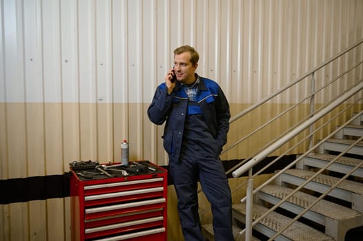 Portrait of a handsome Caucasian man, technician, garage mechanic talking on mobile phone standing near a box with work tools for inspect and repair automobiles and car service station