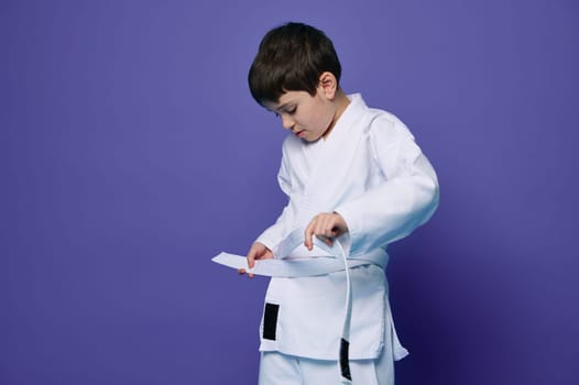 Confident European teenage boy puts on white kimono, ties a belt around his waist, ready for combat sports. Aikido fighter. Oriental martial arts
