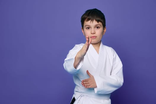 Martial arts attack. School age boy aikido fighter in white kimono isolated over purple background with copy space for advertising text.