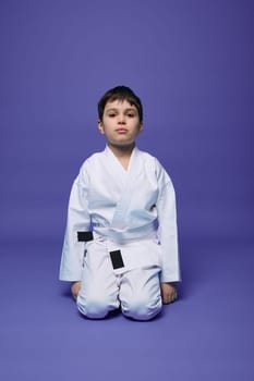 Portrait of a handsome child boy wearing kimono and sitting in aikido stance against purple background with copy space for text. Oriental martial arts concept