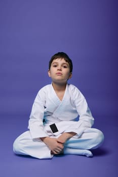Confident European teenager - aikido fighter - in white kimono sitting in lotus pose while practicing oriental martial arts on purple background with copy space