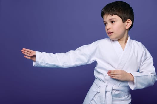 Aikido wrestler European 10 years old boy in white kimono improves his fighting skills, isolated on purple background copy space. Oriental martial arts.