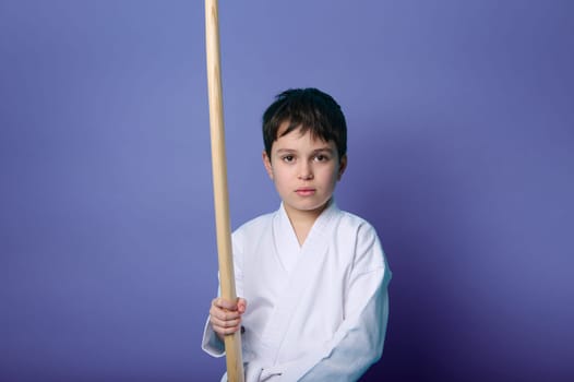 Waist length portrait of charming tranquil confident child boy aikido wrestler holding a wooden sword isolated on purple background with copy ad space