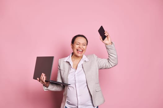 Pregnant middle-aged business woman expressing excitement and joy over pink background, holding laptop and mobile phone