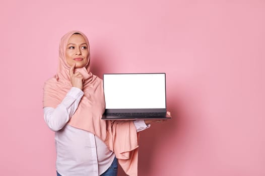 Pensive working pregnant Muslim woman in pink hijab, holds laptop with white screen, reasoning over her maternity leave