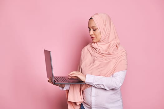 Multitasking Arab Muslim pregnant woman in pink hijab, typing text on laptop keyboard. Pregnancy, business and career