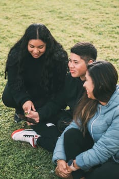 three people of hispanic-latino ethnicity sitting on the green grass of a park, with smart device