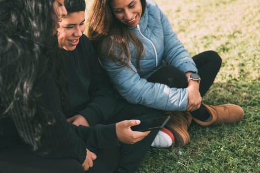 Hispanic male teenager holding smartphone looking away while sitting on grass with Hispanic mother and sister in park on sunny day