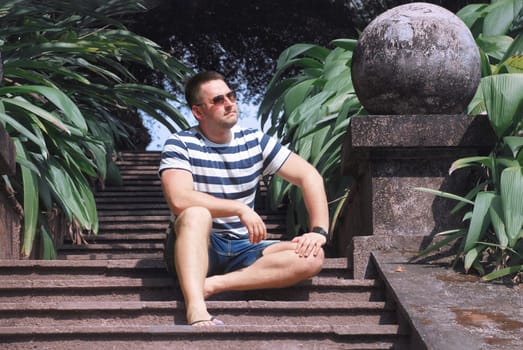 a stylish guy with glasses is sitting on the steps in a tropical garden