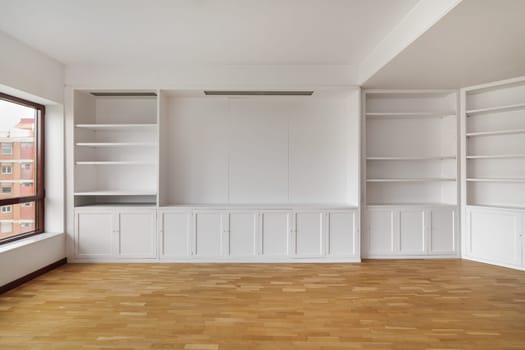 Empty built-in white cabinet on wooden laminate floor with big window in empty bright airy room in housewarming. Concept of housewarming and convenient storage