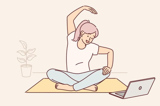 Woman sits on yoga mat and looks at laptop screen doing exercises during online broadcast