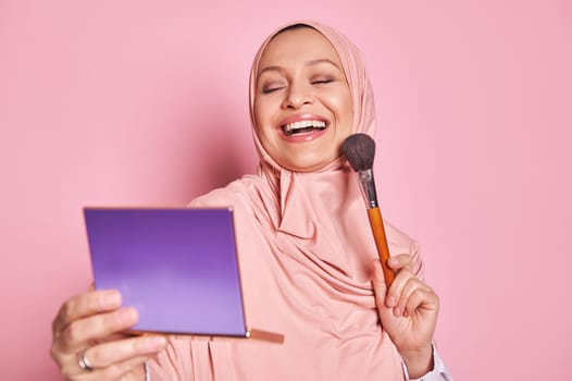 Smiling Muslim woman in pink hijab, applying foundation powder or blush with makeup brush on her cheek, isolated on pink
