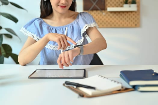 Cropped shot of young woman checking time on her smartwatch and using digital tablet at working desk