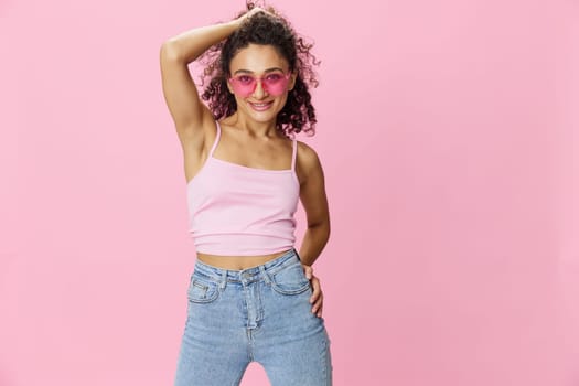 Happy woman hands up waxing her armpits, shaving, with curly hair in a pink tank top and jeans on a pink background wearing sunglasses with a nice tan, copy space