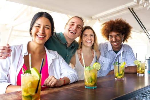 Happy multiracial friends enjoying cocktails at beach bar and having fun looking at camera.