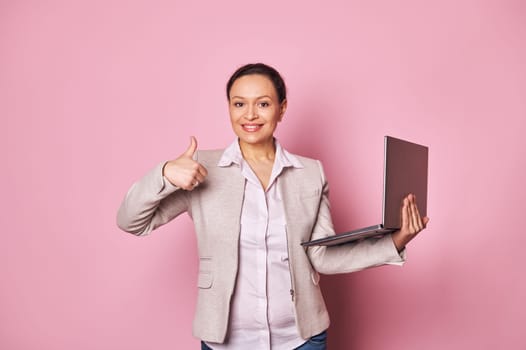 Confident multi ethnic woman sales manager showing thumb up, holding laptop, smiling looking at camera, isolated on pink