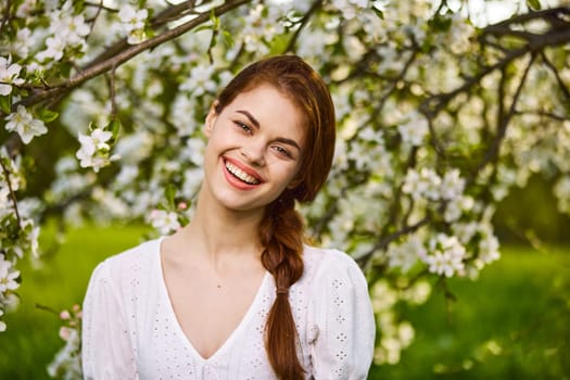 a joyful, carefree woman in a light dress stands against the background of a flowering tree