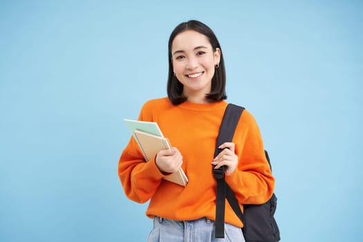 Education and students. Smiling young asian woman with backpack and notebooks, posing against blue background
