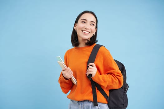 Education and students. Smiling young asian woman with backpack and notebooks, posing against blue background