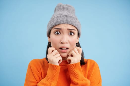 Portrait of asian girl in warm hat, looks with fear, shaking and trembling scared, standing frightened against blue background