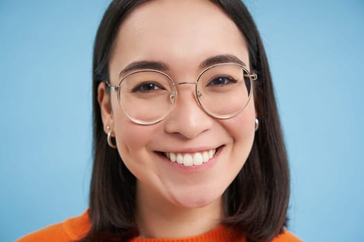 Close up portrait of beautiful young asian woman in glasses, smiling and looking happy, trying new eyewear at opticians store, stands over blue background