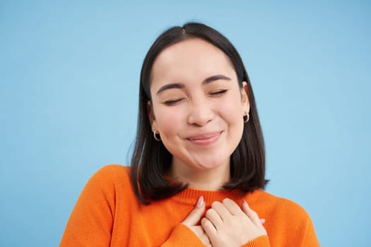 Close up of lovely asian woman, holds hands on heart, daydreaming, thinking of something happy, standing over blue background
