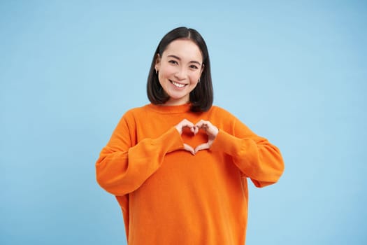 Lovely asian woman, korean girl shows heart gesture and smiles with care and tenderness, stands in orange sweatshirt against blue background