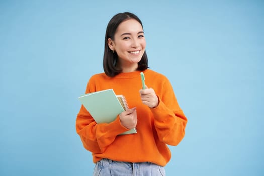 Education and students. Happy asian woman, holding notebooks and laughing, smiling at camera, enjoys going to University or College, blue background