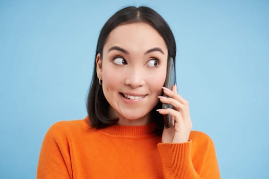 Technology concept. Close up of smiling asian woman talks on mobile phone, having conversation on cellphone, blue background.