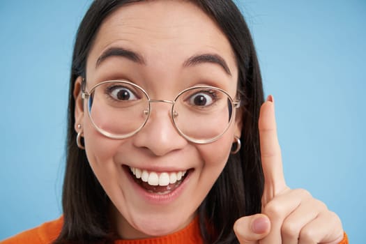 Eureka. Close up portrait of excited korean woman in glasses, shows one finger, has an idea, stands over blue background